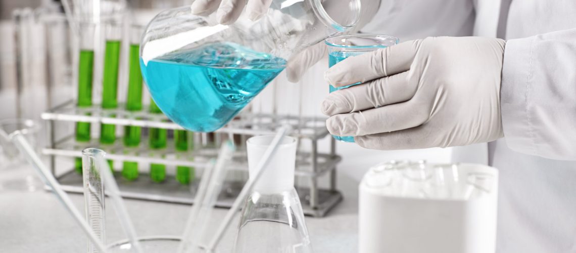 Young clinical worker dressed in white gown and gloves holding glass beakers with blue liquid making experiment in laboratory. Scientific laboratory research, laboratory beakers in hadns of scientist